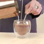 A person in a navy pajama shirt pours milk from a copper pitcher into a clear double-walled glass mug with 'Dreams' printed on it, set on a light wood surface.