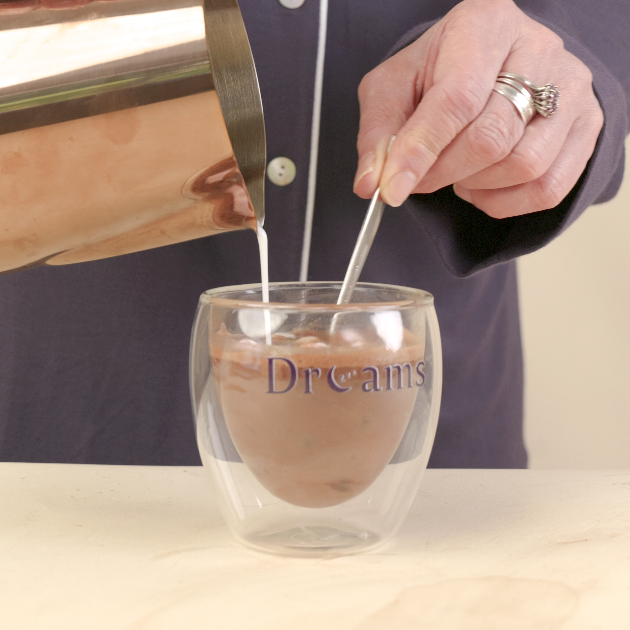 A person in a navy pajama shirt pours milk from a copper pitcher into a clear double-walled glass mug with 'Dreams' printed on it, set on a light wood surface.