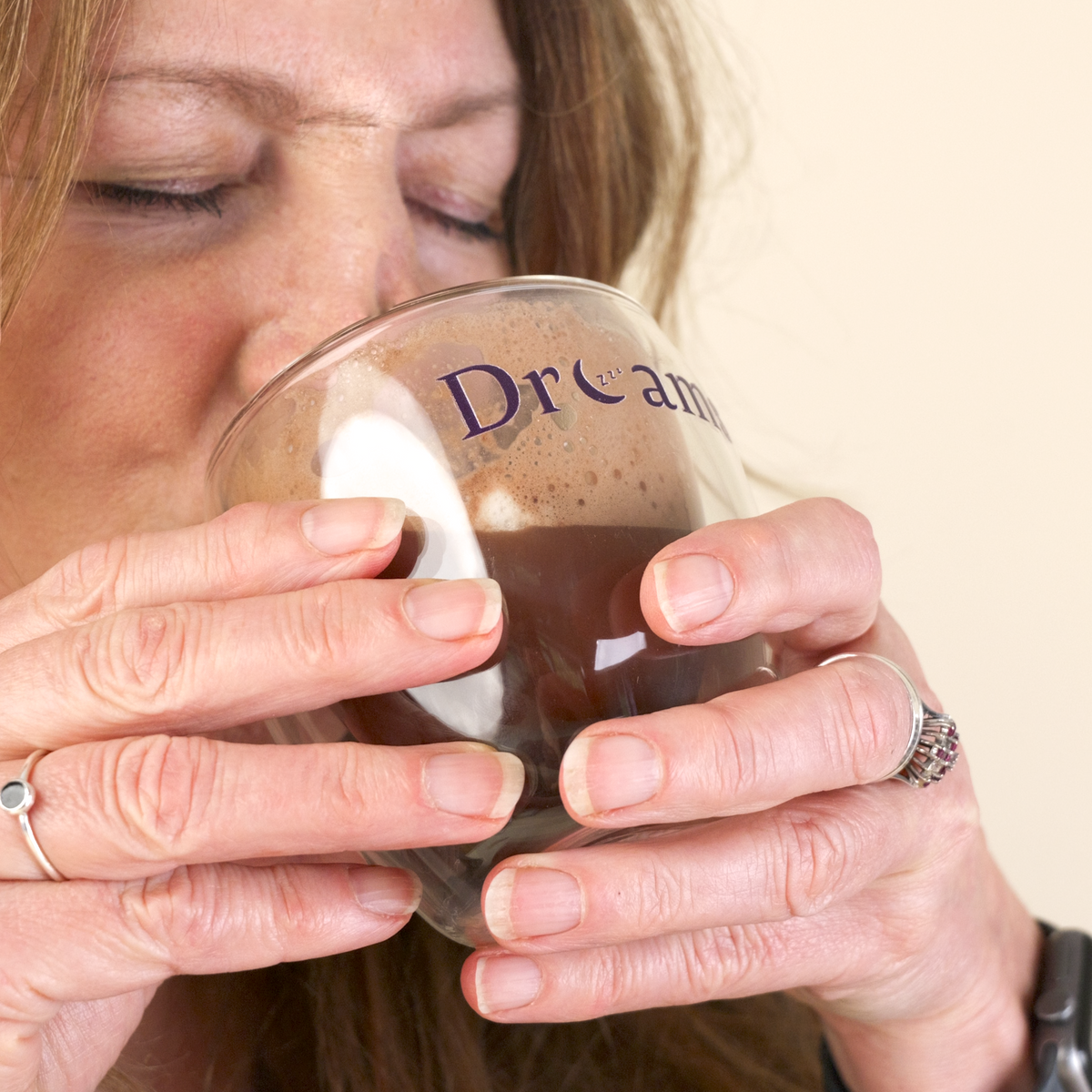 A close-up lifestyle shot of a woman drinking from a clear glass mug with the word 'Dream' printed on it, wearing a dark navy pajama top against a solid beige background.