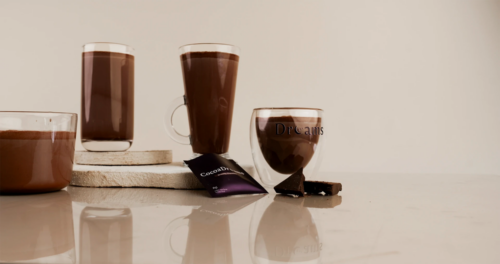 Two glasses of chocolate hot cocoa and a glass of milk chocolate with chocolate pieces on a reflective white surface, alongside a chocolate sachet and a glass cup with 'Dreams' printed on it, set against a warm beige background.