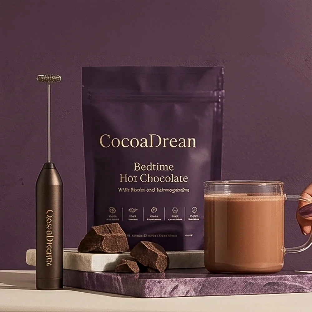CocoaDreams bedtime hot chocolate pouch with frother, chocolate pieces, and a glass mug on a purple backdrop