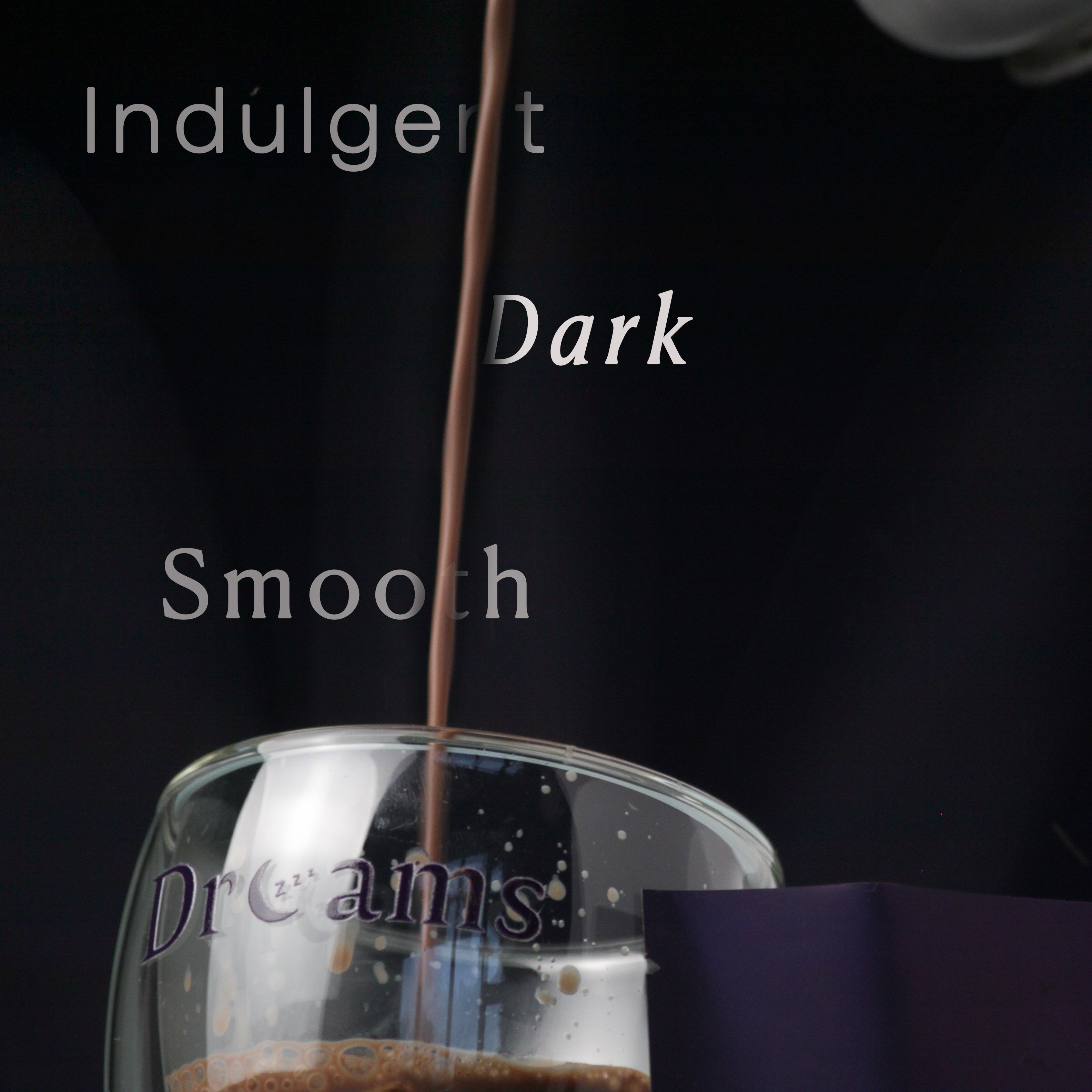 Glass of dark chocolate drink with a bar of chocolate and a box labeled 'Cocoa Dreams' on a dark background.