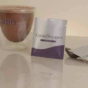 A double-walled clear glass cup filled with cocoa drink sits on a light beige surface next to a standing and a lying flat packet of CocoaDreams powder. The background is a plain beige wall.